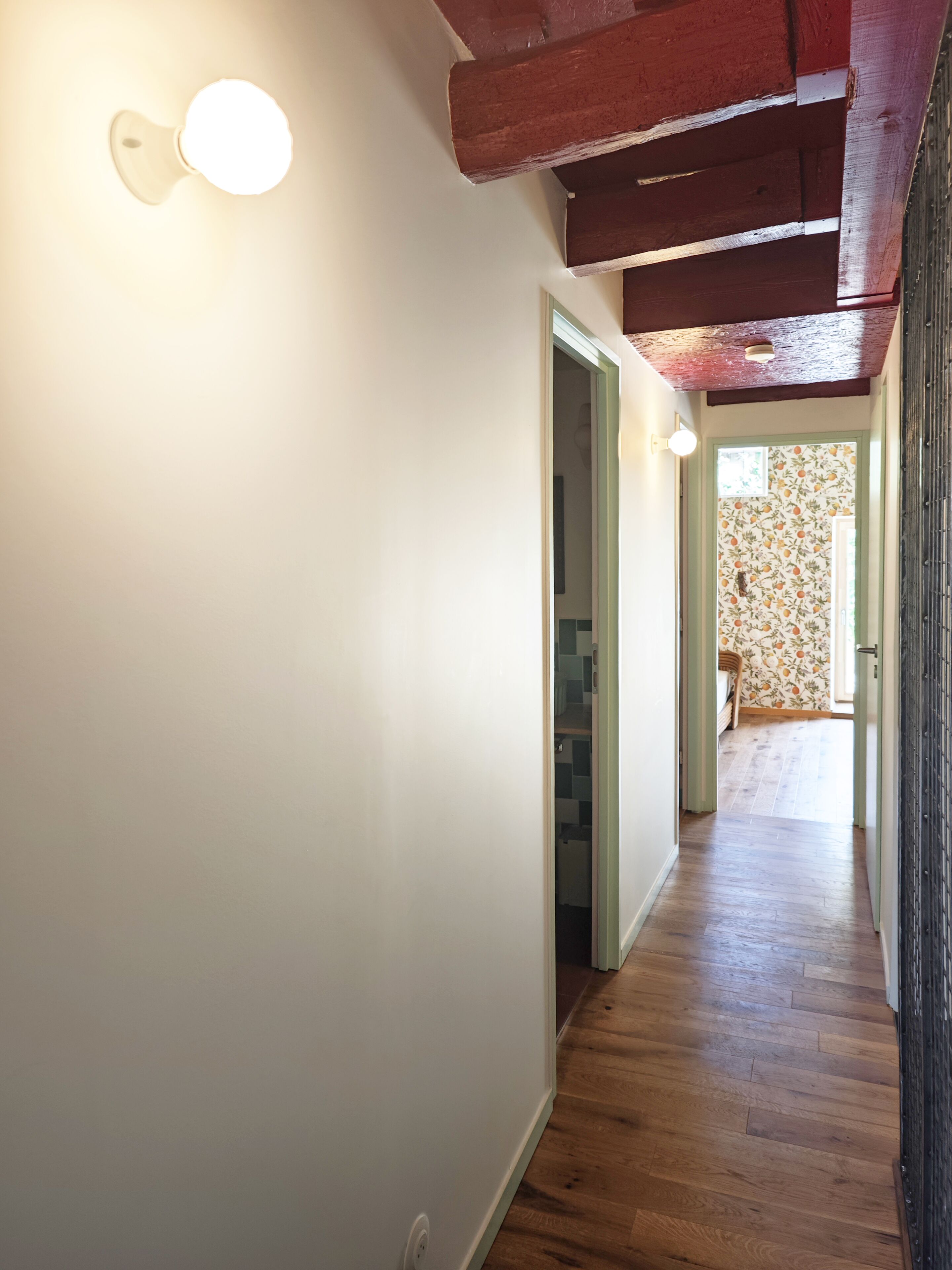 A hallway with wooden flooring, a light fixture on the wall, and a door leading to another room.