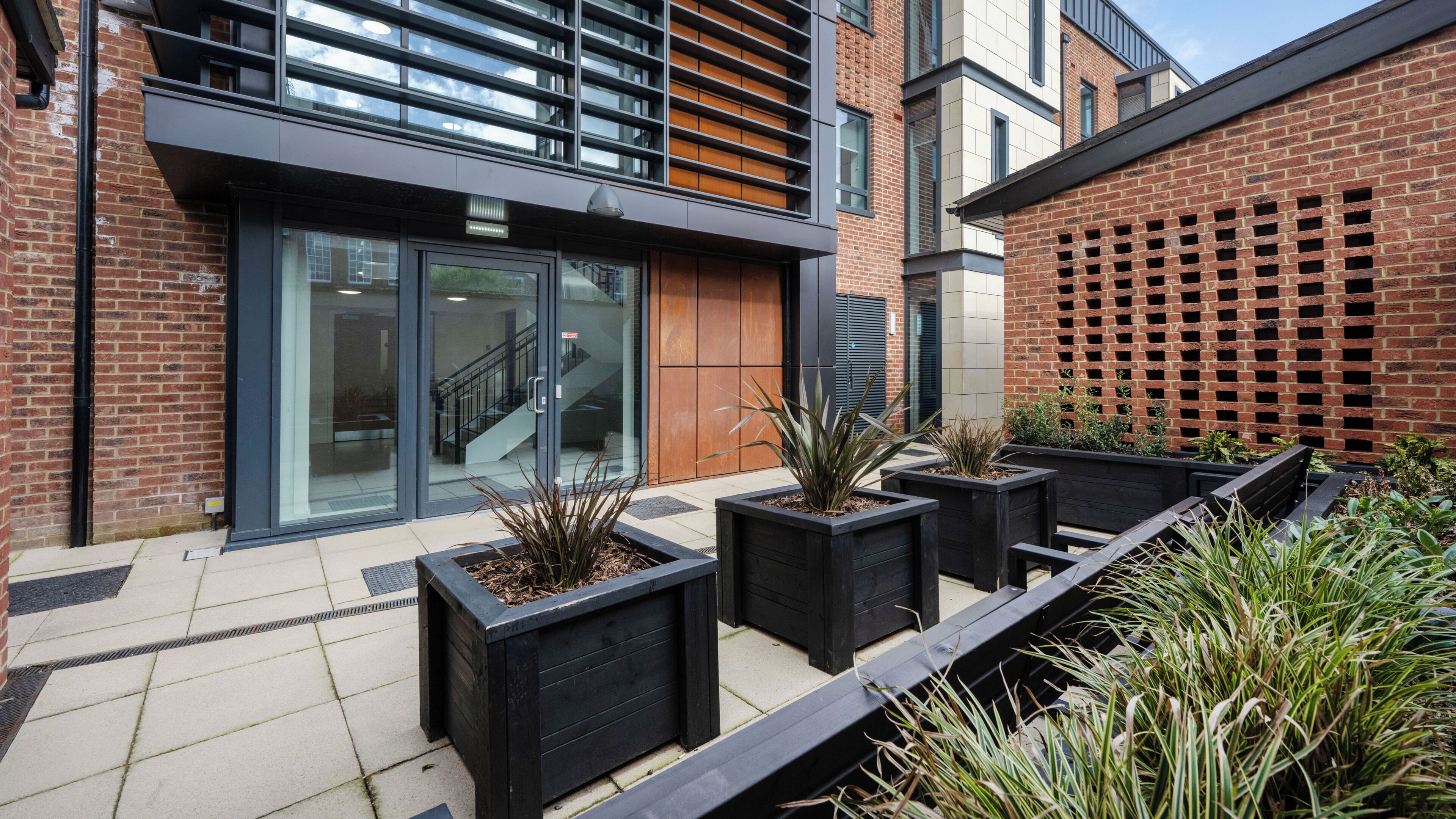 A modern building with a glass entrance, a wooden door, and a brick wall with a lattice design. There are black planters with plants and a bench.