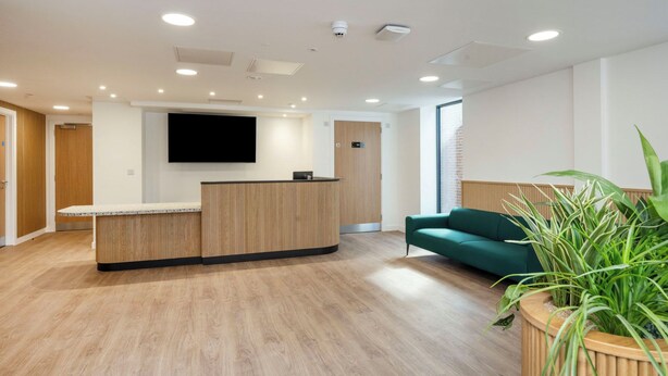 A reception area with a wooden reception desk, a flat-screen TV, a green sofa, and a potted plant. - Student Only Waterloo House (York)