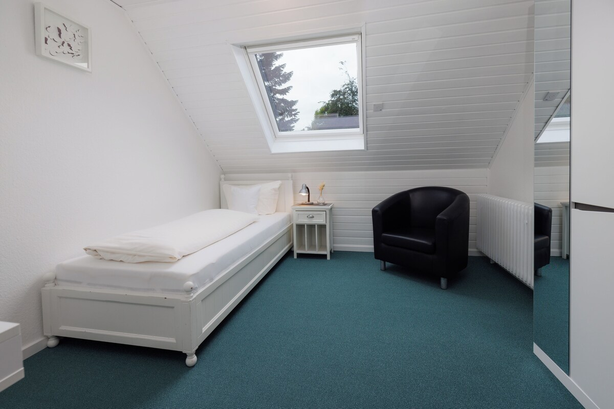 Een kleine, witte eenpersoonskamer met dakraam, een zwarte stoel en een radiator.