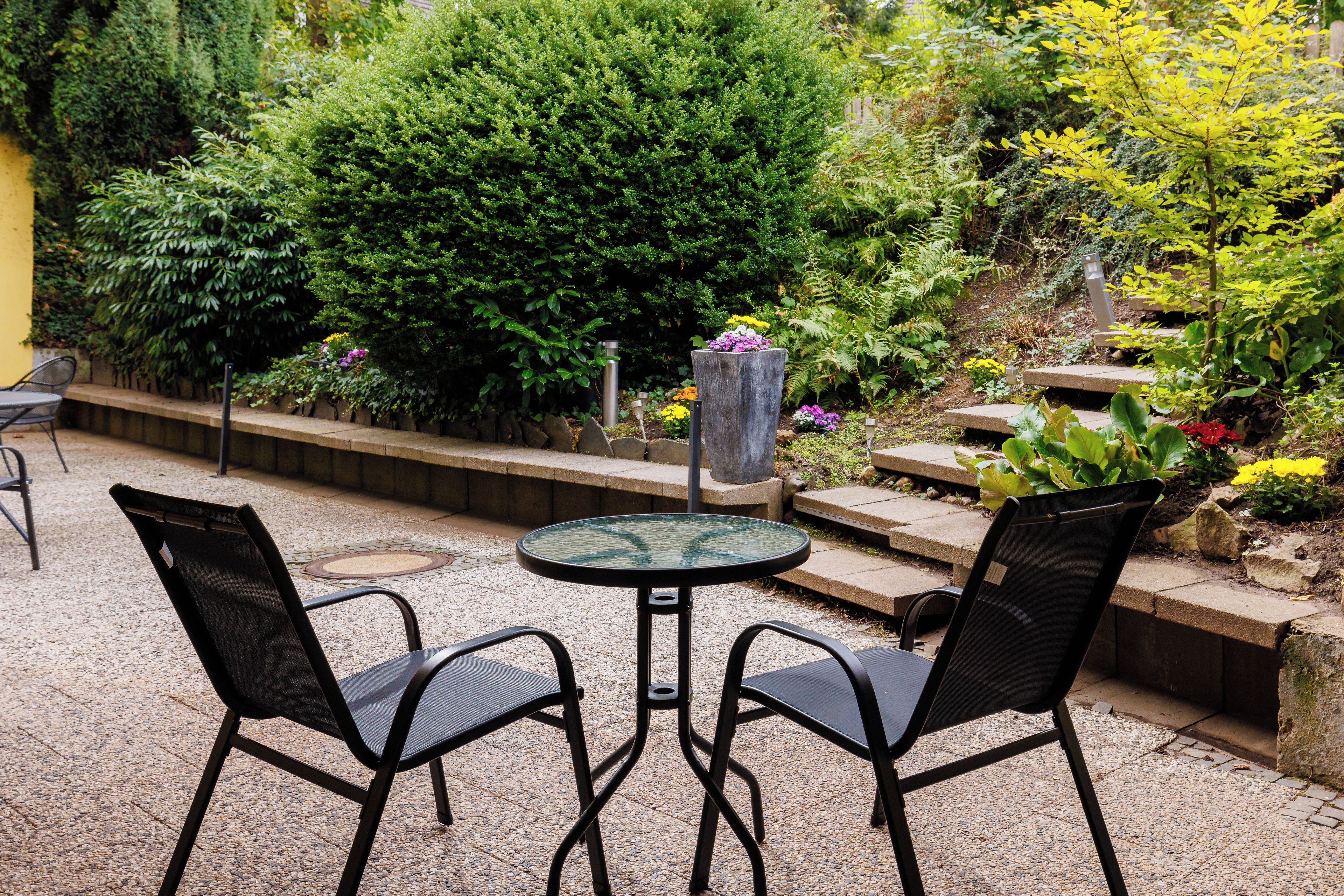 Een terras met een ronde tafel en twee stoelen, omgeven door verschillende planten en een stenen pad.
