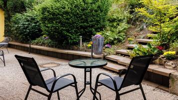 Een terras met een ronde tafel en twee stoelen, omgeven door verschillende planten en een stenen pad.