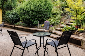 An outdoor seating area with a round table and two chairs, surrounded by a variety of plants and a stone pathway.