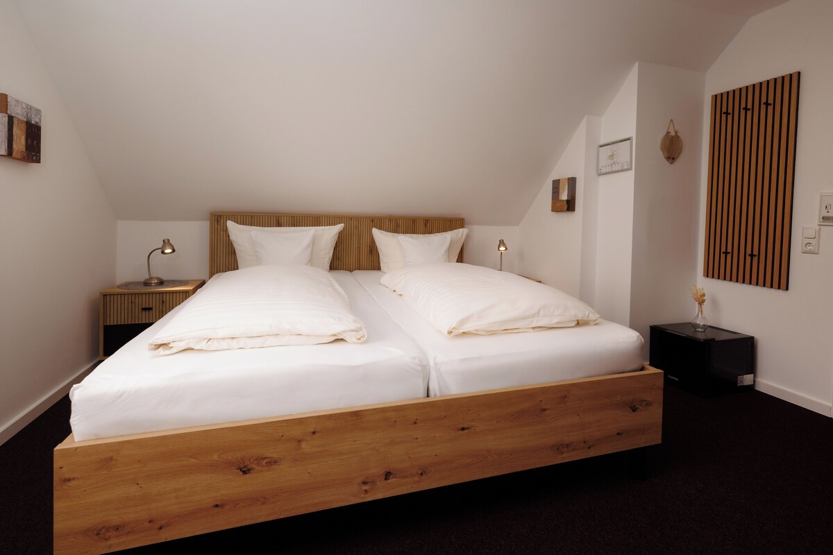 Een tweepersoonsbed met houten frame, twee nachtkastjes met lampen, een houten paneel aan de muur en een klein zwart object op de vloer.