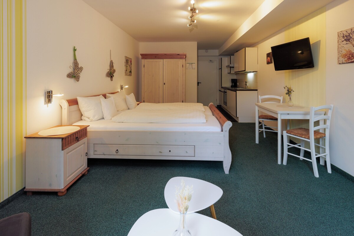 Een slaapkamer met een bed, nachtkastje, kaptafel, een klein rond tafeltje met een vaas, een aan de muur bevestigde televisie, en een keuken in de achtergrond.
