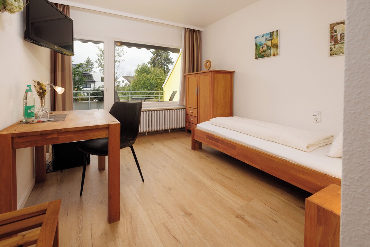 Een kleine, goed verlichte kamer met een houten bureau, een stoel, een bed en een radiator.