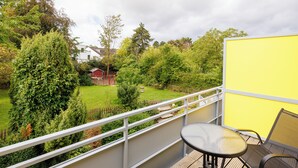 Ein Balkon mit Tisch und Stühlen, der auf einen Garten mit verschiedenen Bäumen und einem roten Gebäude im Hintergrund blickt.