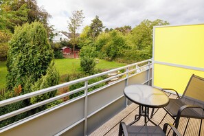 Een balkon met een tafel en stoelen, met uitzicht op een tuin met verschillende bomen en een rood gebouw op de achtergrond.