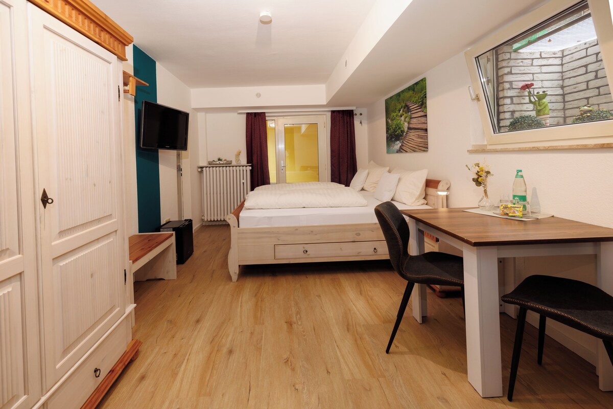 Een slaapkamer met een bed, een bureau, een stoel, een radiator, een televisie en een raam met uitzicht op een gebouw.