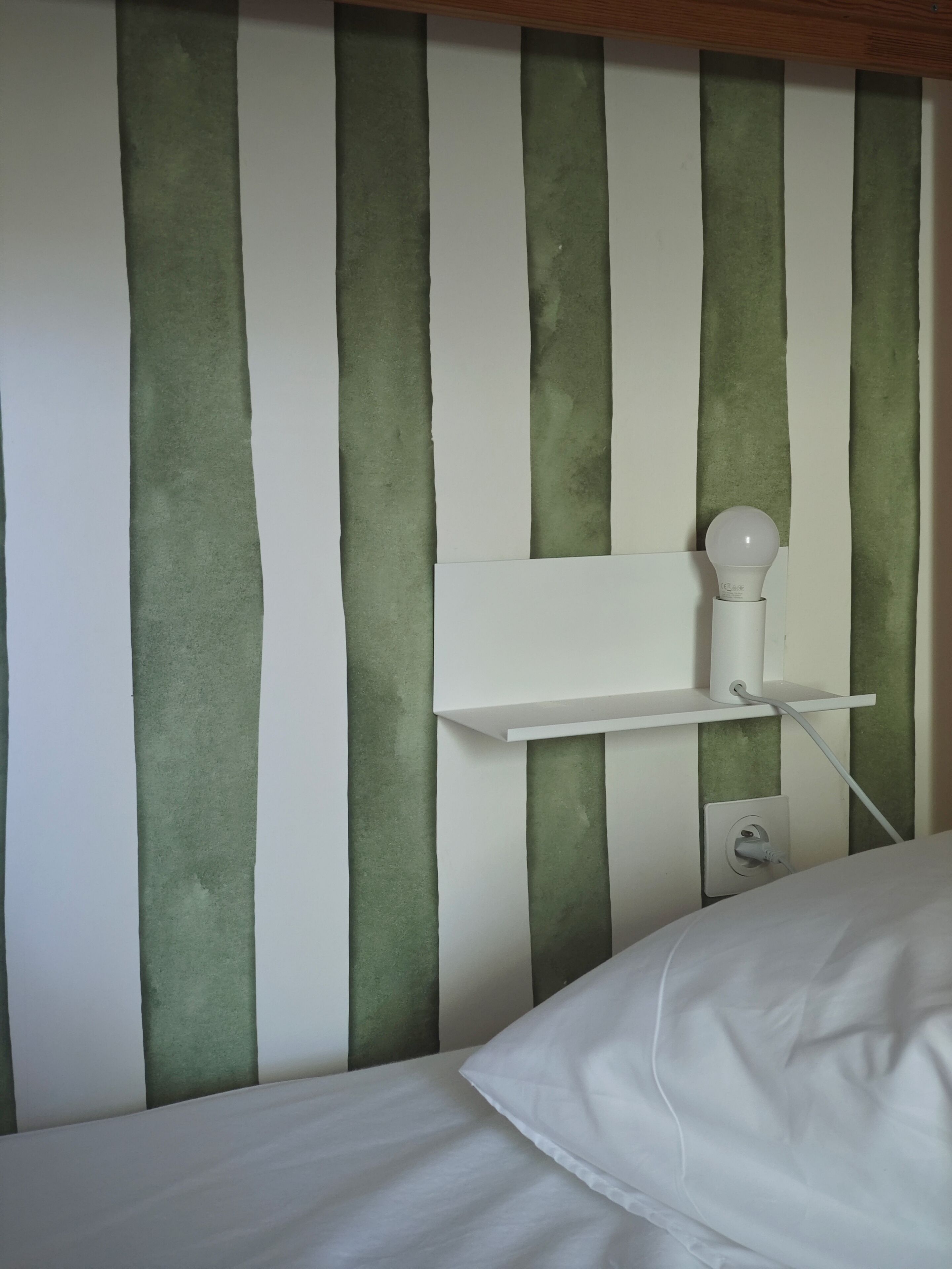 A bed with white bedding, a white bedside table with a lamp, and a wall with vertical green and white stripes.