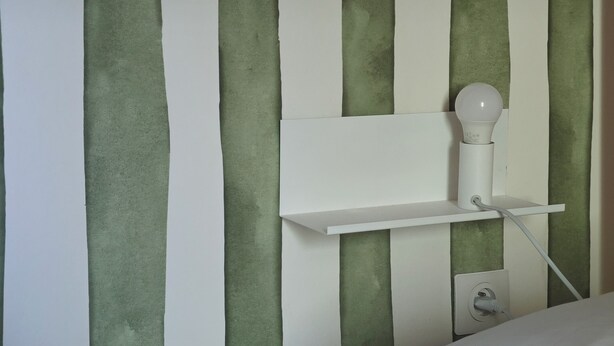 Ein Bett mit weißer Bettwäsche, ein Nachttisch in Weiß mit einer Lampe sowie eine Wand mit senkrechten, grünen und weißen Streifen.