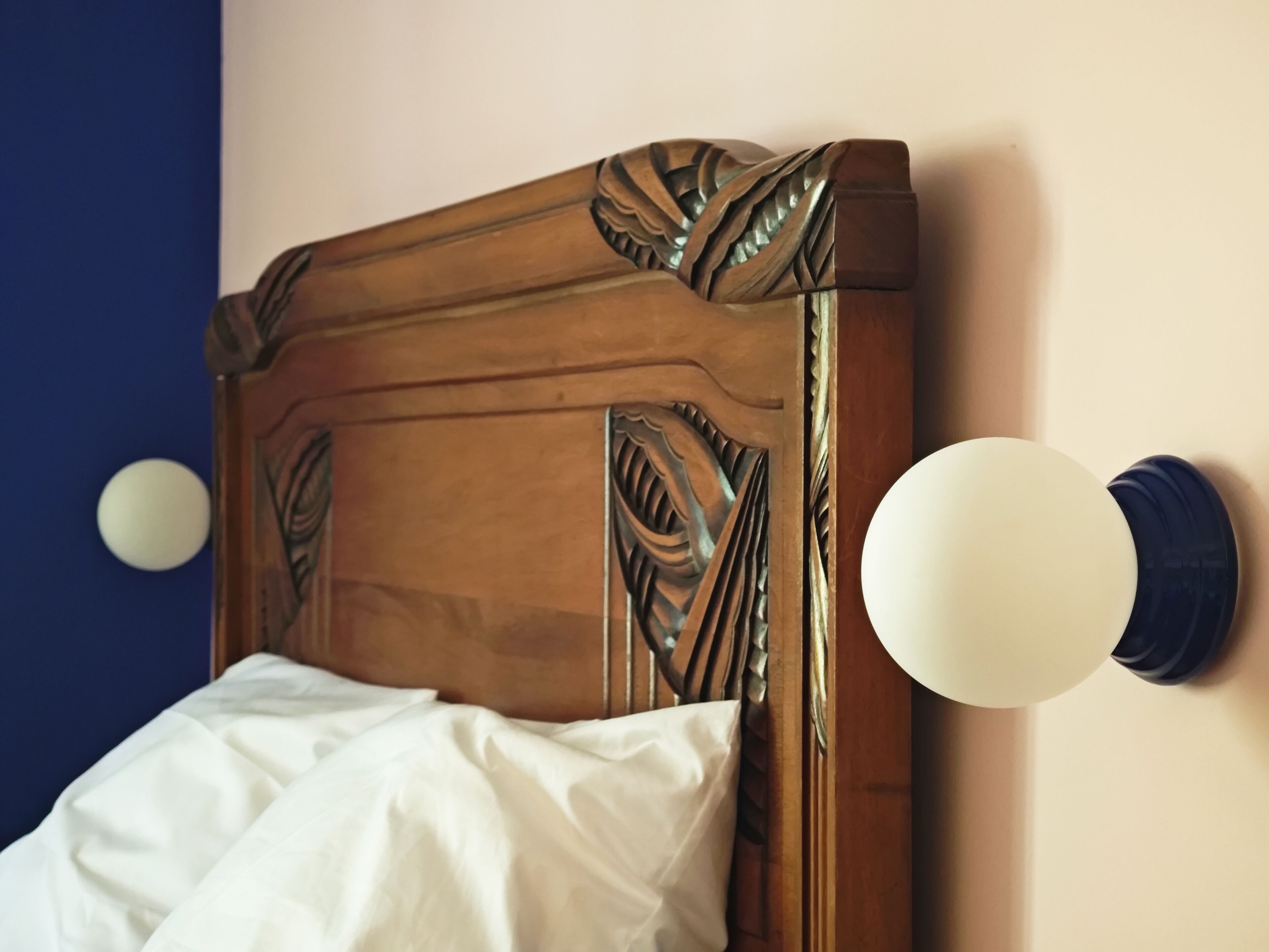 Een bed met een houten hoofdbord met ingewikkelde houtsnijwerken, een wit kussen en een rond, aan de muur bevestigd lampje.