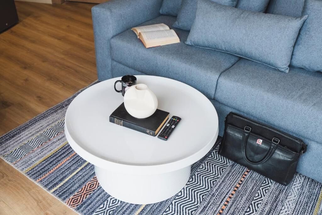 A round white coffee table with a white teapot, a book, and a remote control. A black handbag is placed beside the table on a patterned rug. A blue sofa with a book on it is in the background.