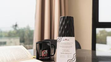 Uma caneca preta com um logotipo, um vaso preto e um livreto sobre uma mesa com vista para a janela.
