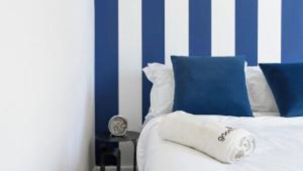 A bed with white bedding and blue pillows, a bedside table with a clock, and a wall with vertical blue and white stripes.