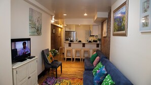Ein Wohnzimmer mit Fernseher, einer Couch mit gemusterten Kissen, einem Couchtisch und einer Küche im Hintergrund.