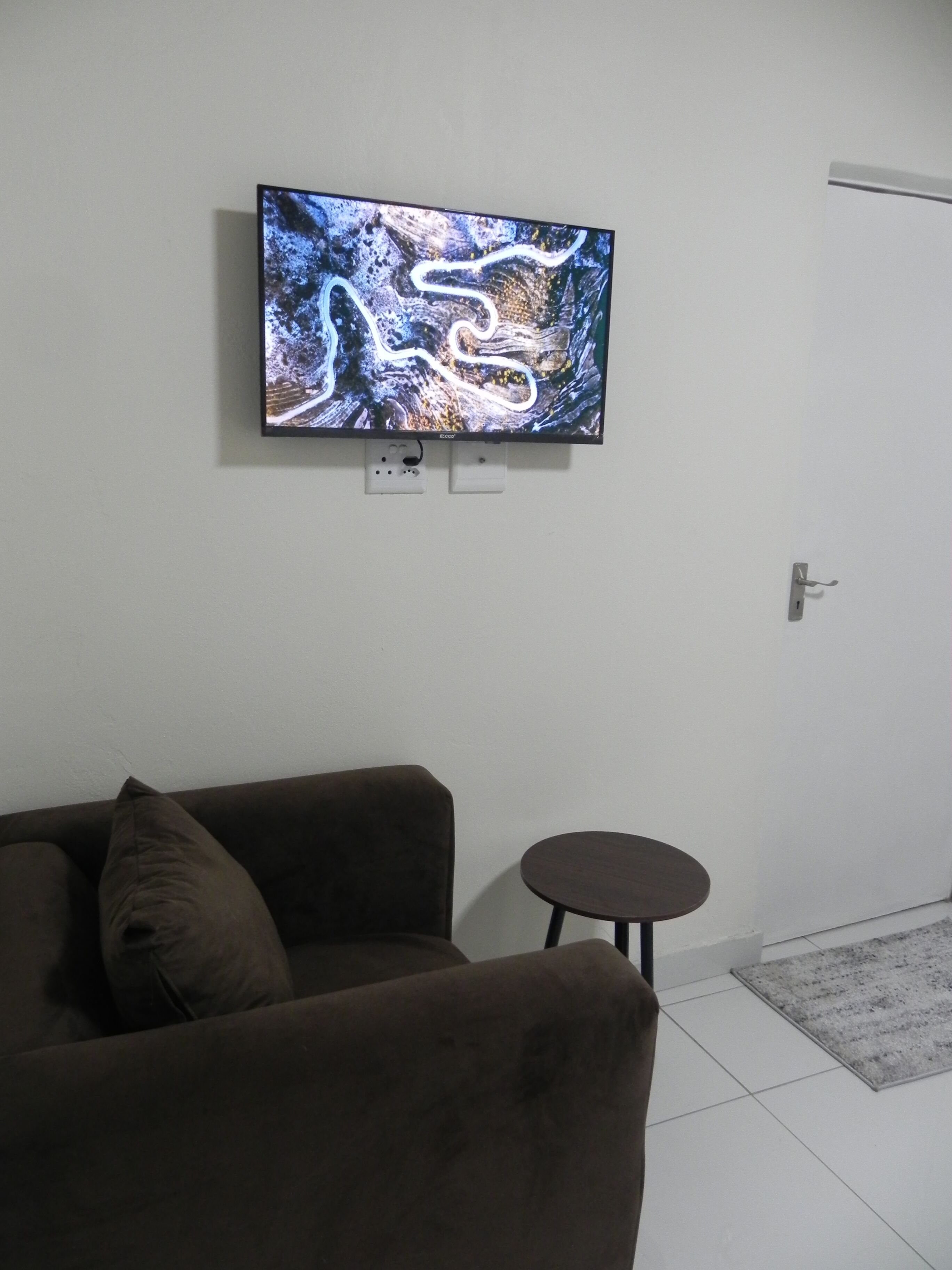 A living room with a flat-screen TV mounted on the wall, a brown sofa, a small round table, and a door.