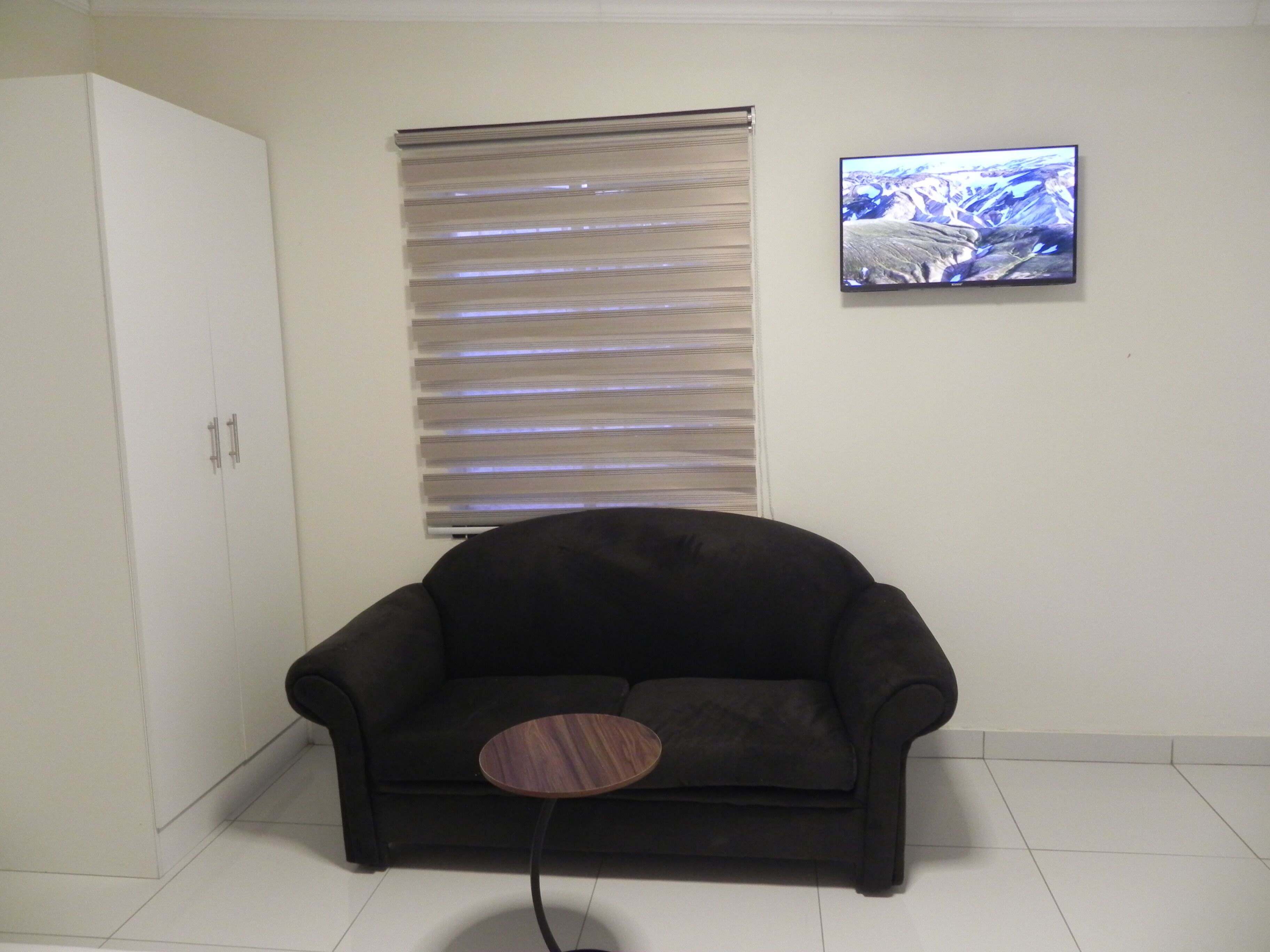A room with a black sofa, a small round table, a white wardrobe, a window with blinds, and a framed picture on the wall.