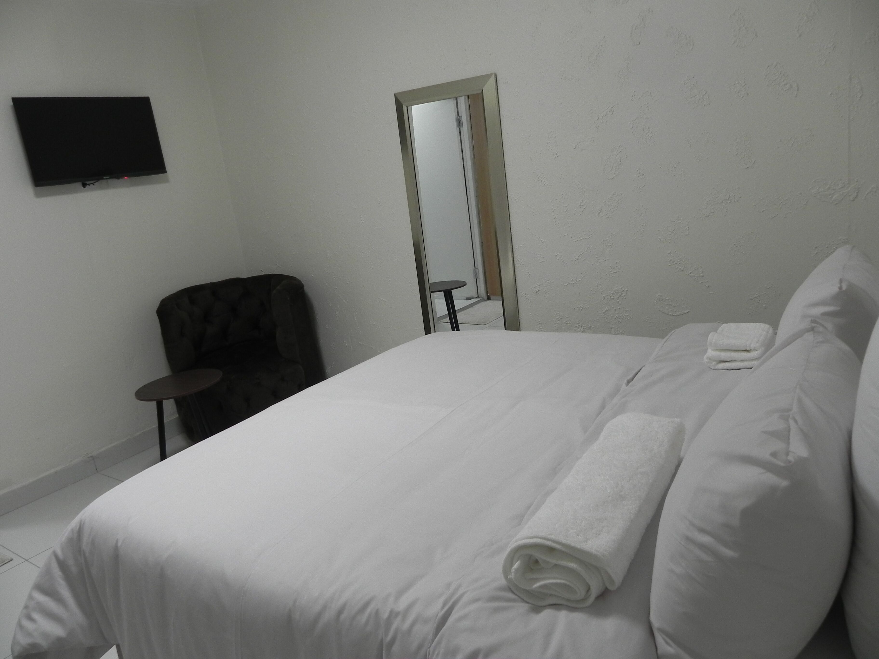 A hotel room with a bed, a chair, a small table, a flat-screen TV mounted on the wall, and a mirror.