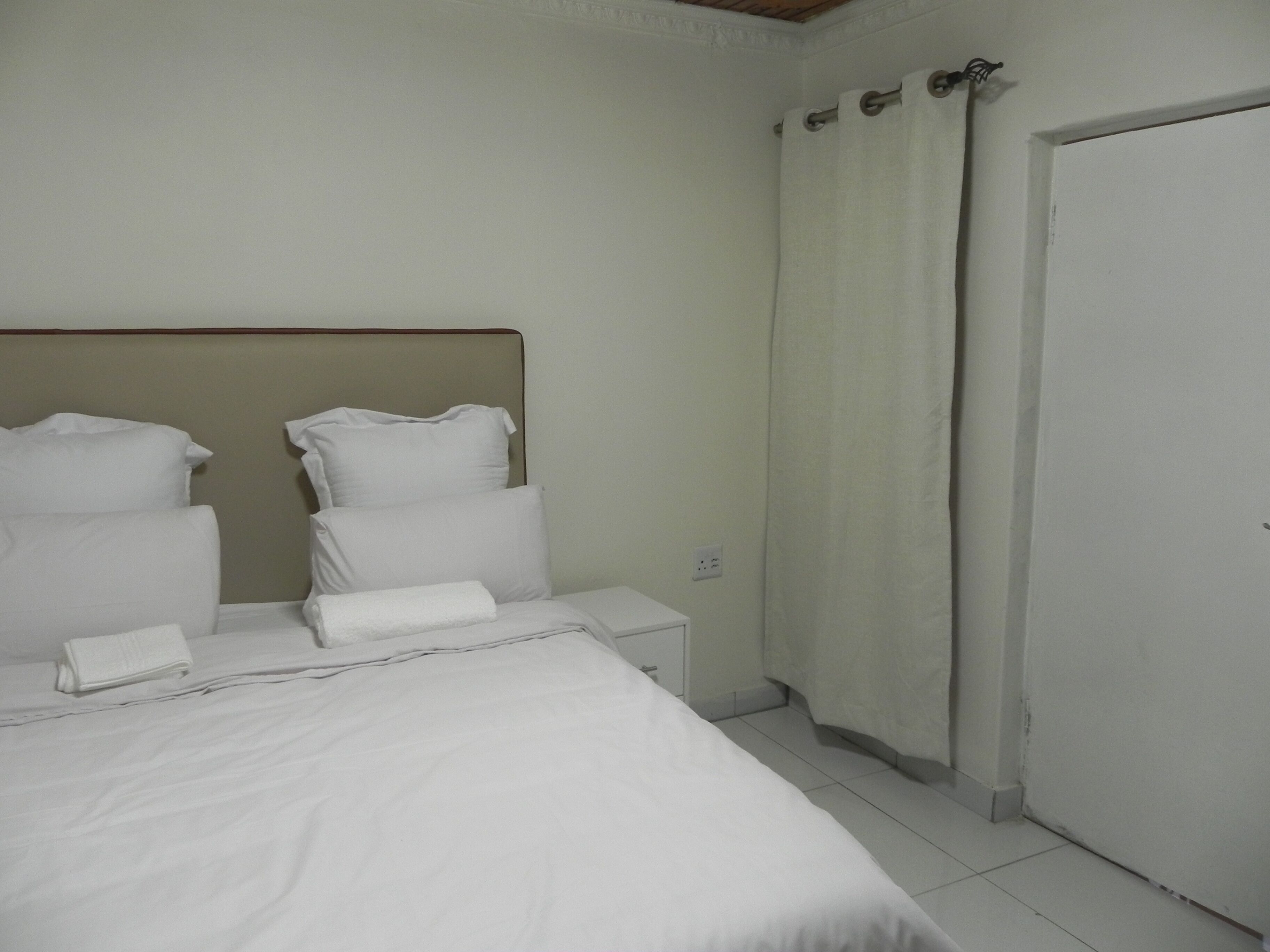 A hotel room with a bed, pillows, a nightstand, a curtain, and a door.