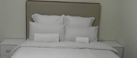 Una cama bien hecha con sábanas y almohadas blancas, un control remoto sobre la cama y una pared lisa al fondo.