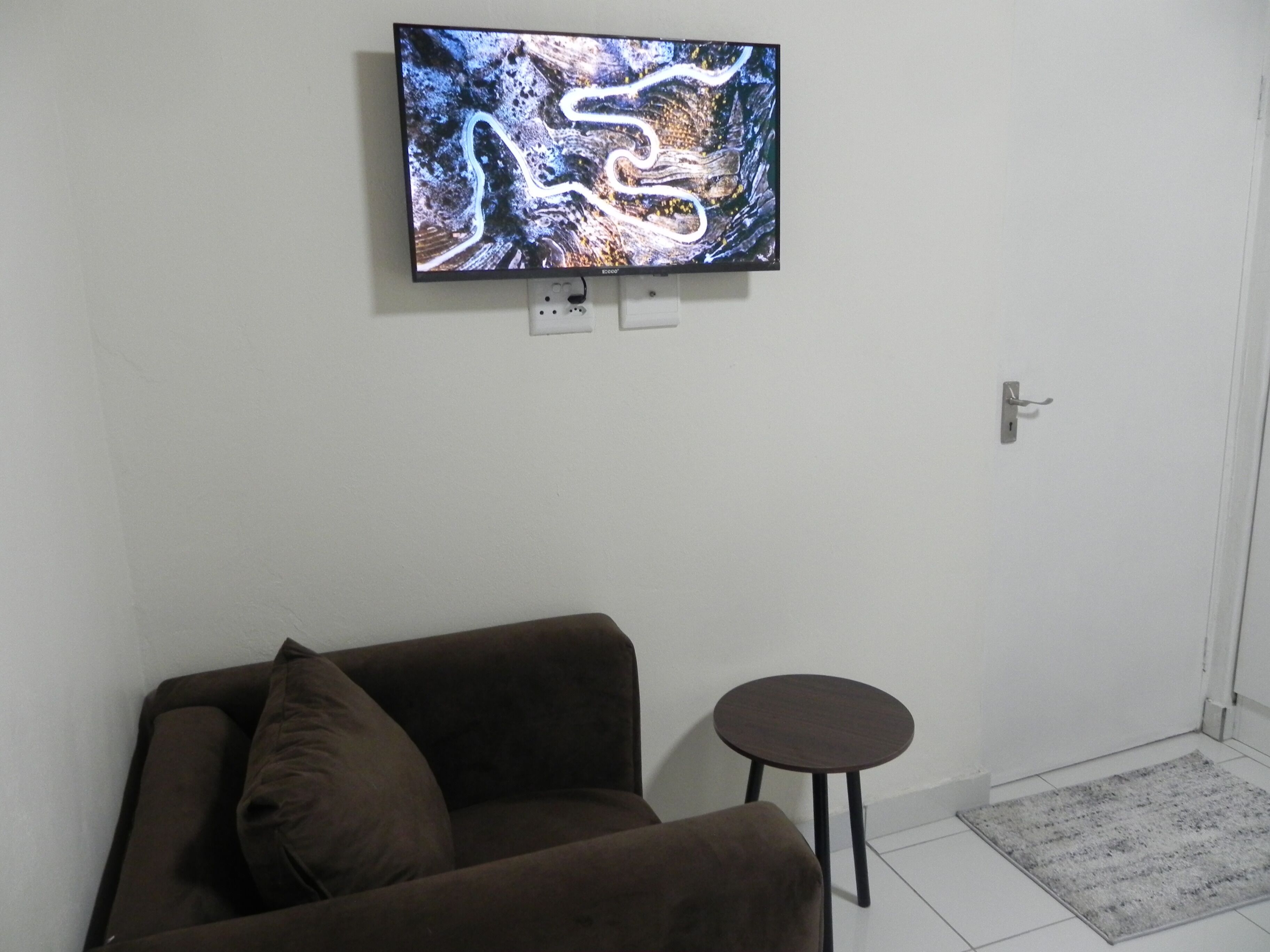 A room with a brown sofa, a small round table, and a flat-screen TV mounted on the wall.