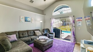 Ein Wohnzimmer mit einer Eckcouch, einer Liegesessel und einem violetten Teppich. Ein großes Fenster mit Vorhängen, ein Pool im Freien und verschiedene Wanddekorationen sind zu sehen.