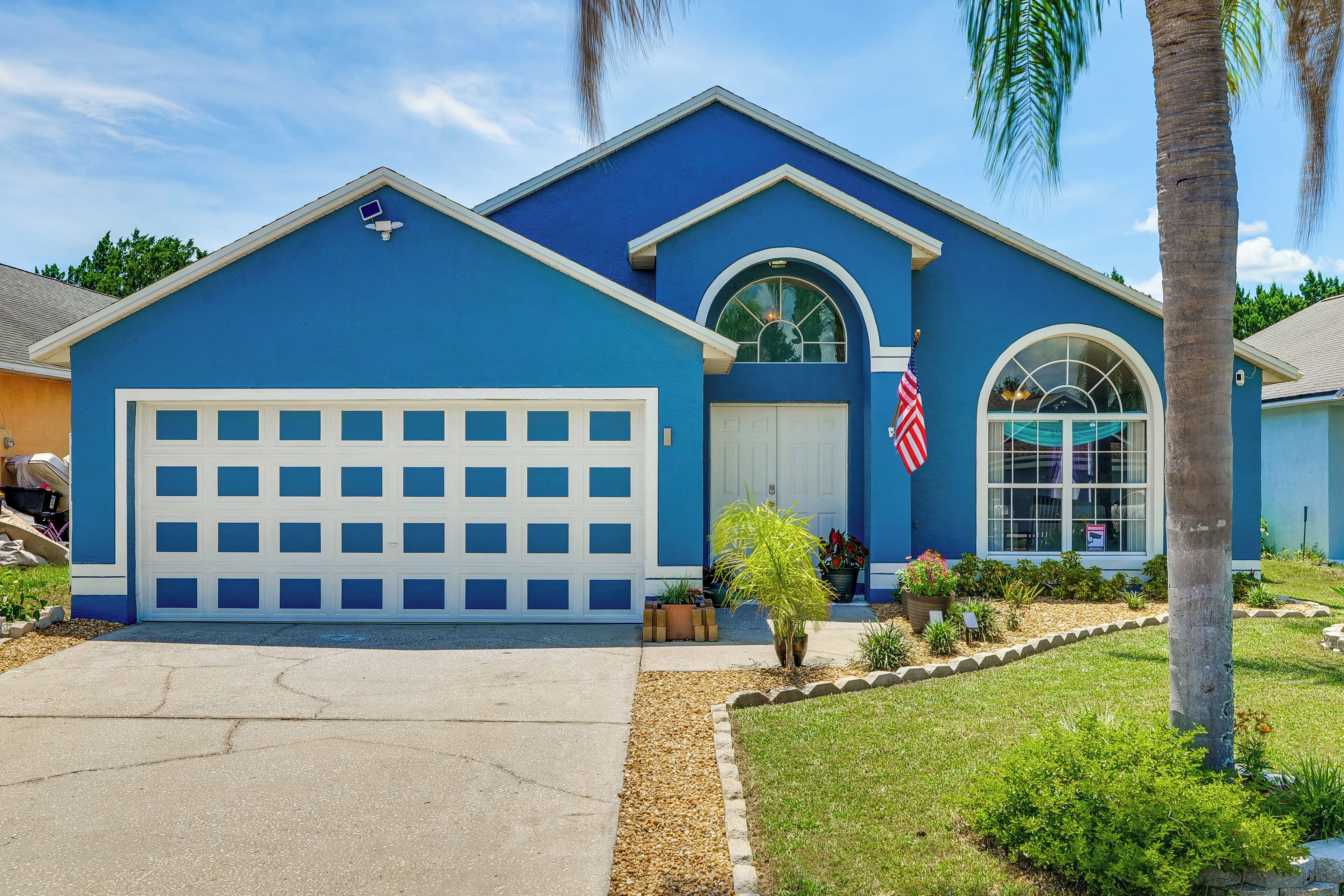 A residential building with a blue exterior, a white garage door, and a small front yard with a palm tree and potted plants.
