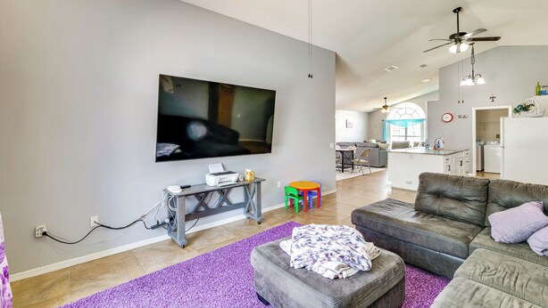 Ein modernes Wohnzimmer mit Flachbildfernseher, grauem Sofa, violettem Teppich und einem Essbereich mit kleinem Tisch und bunten Stühlen.