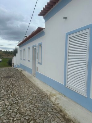 A building with a white exterior and blue trim, a tiled roof, and a stone-paved ground. - Family home in an Alentejo village (Amieira Portel)