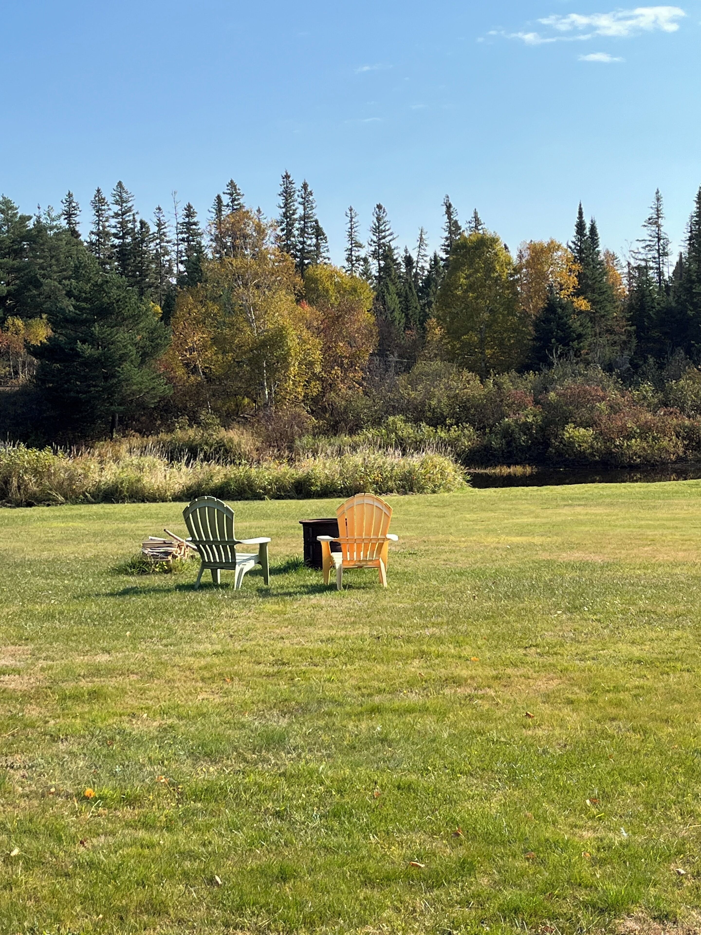 An outdoor seating area with two Adirondack chairs and a small table, surrounded by a grassy field and a forested backdrop.