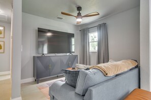A modern living room with a grey sofa, a flat-screen TV mounted on a cabinet, a ceiling fan, and a window with curtains. - 4 Mi to Beach! Dual-unit Safety Harbor Retreat (Safety Harbor)