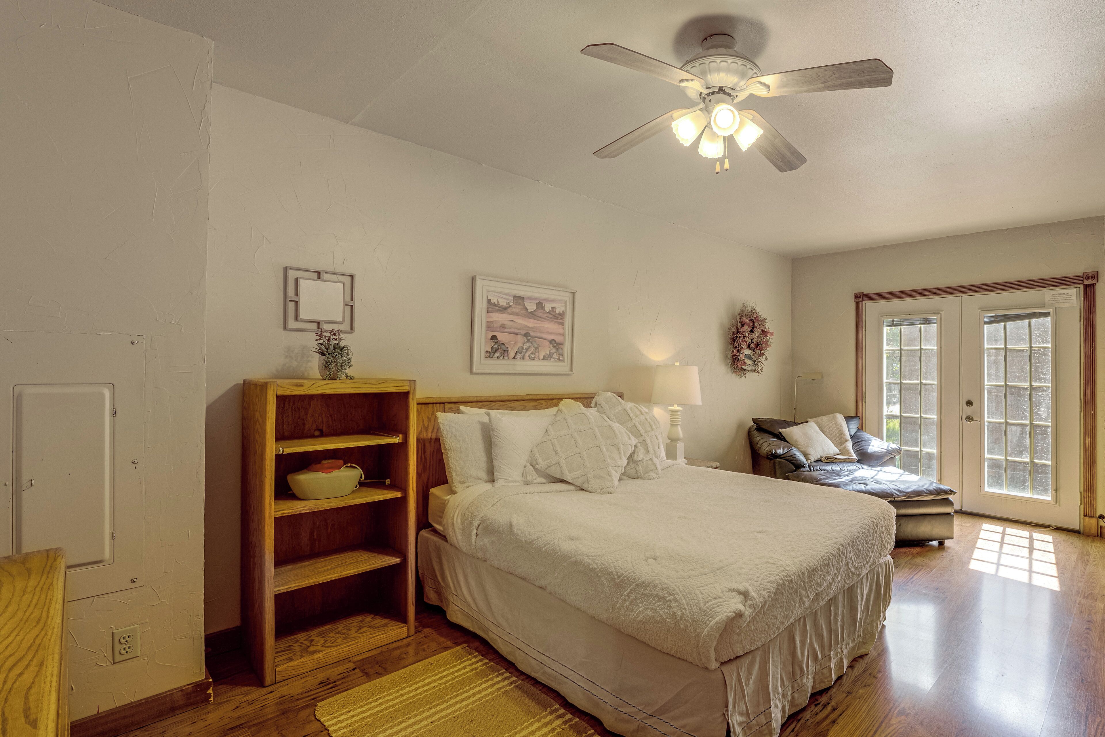 A bedroom with a bed, a wooden shelf, a painting, a ceiling fan, and a window.