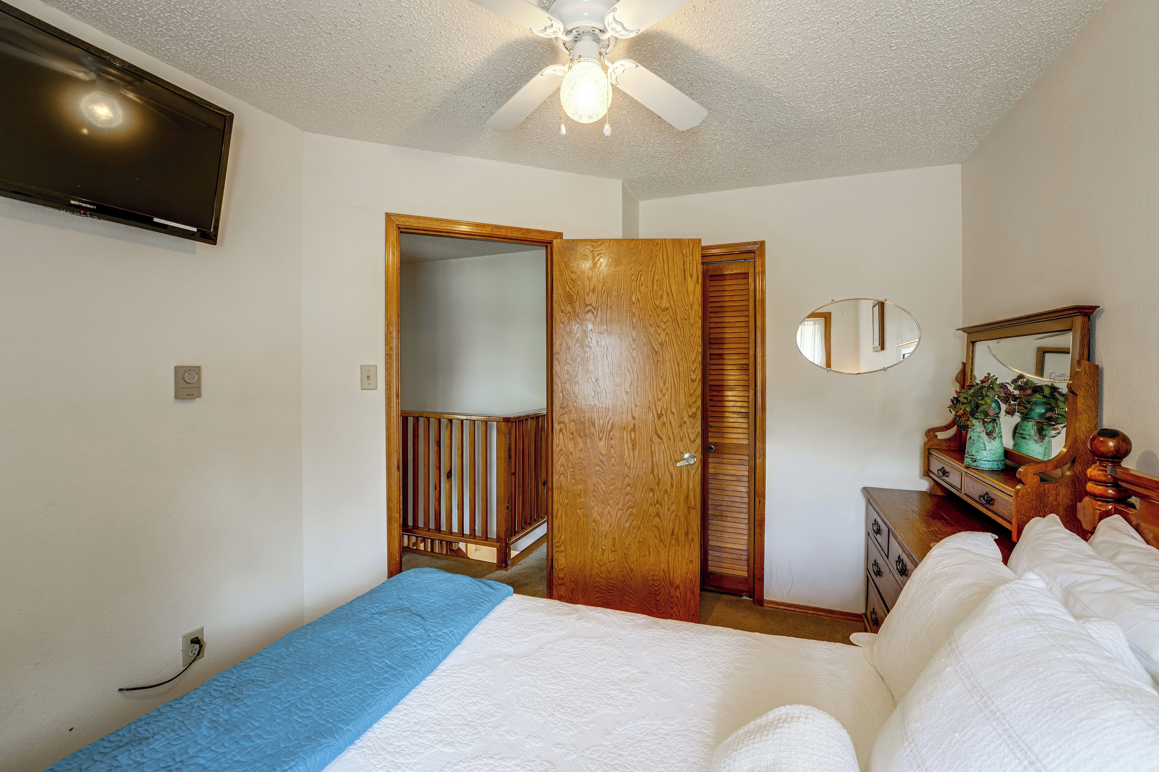 A bedroom with a bed, a ceiling fan, a television mounted on the wall, and a wooden wardrobe.