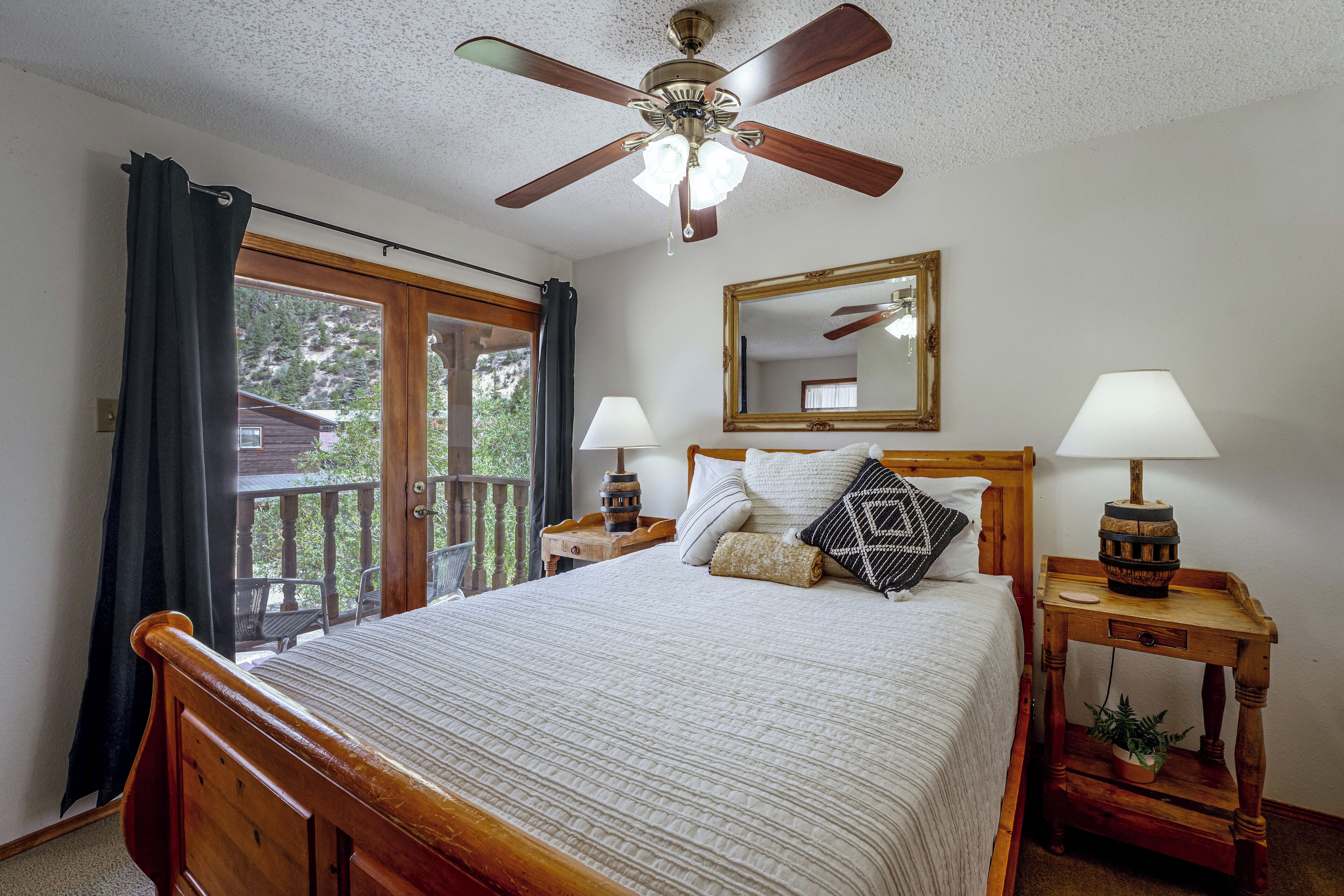 A bedroom with a wooden bed, two bedside tables with lamps, a ceiling fan, a large window with curtains, and a mirror on the wall.