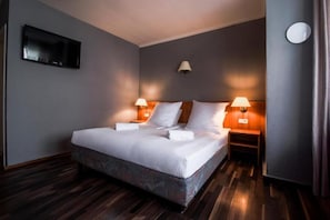 Quarto de hotel com uma cama grande, duas luminárias de cabeceira, uma televisão de tela plana na parede e piso de madeira.