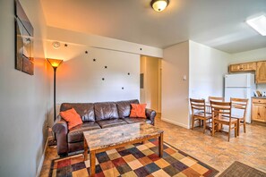 A living room with a brown leather sofa, a coffee table, and a patterned rug. There is a dining area with wooden chairs and a small table. The kitchen is visible with wooden cabinets and a white refrigerator.