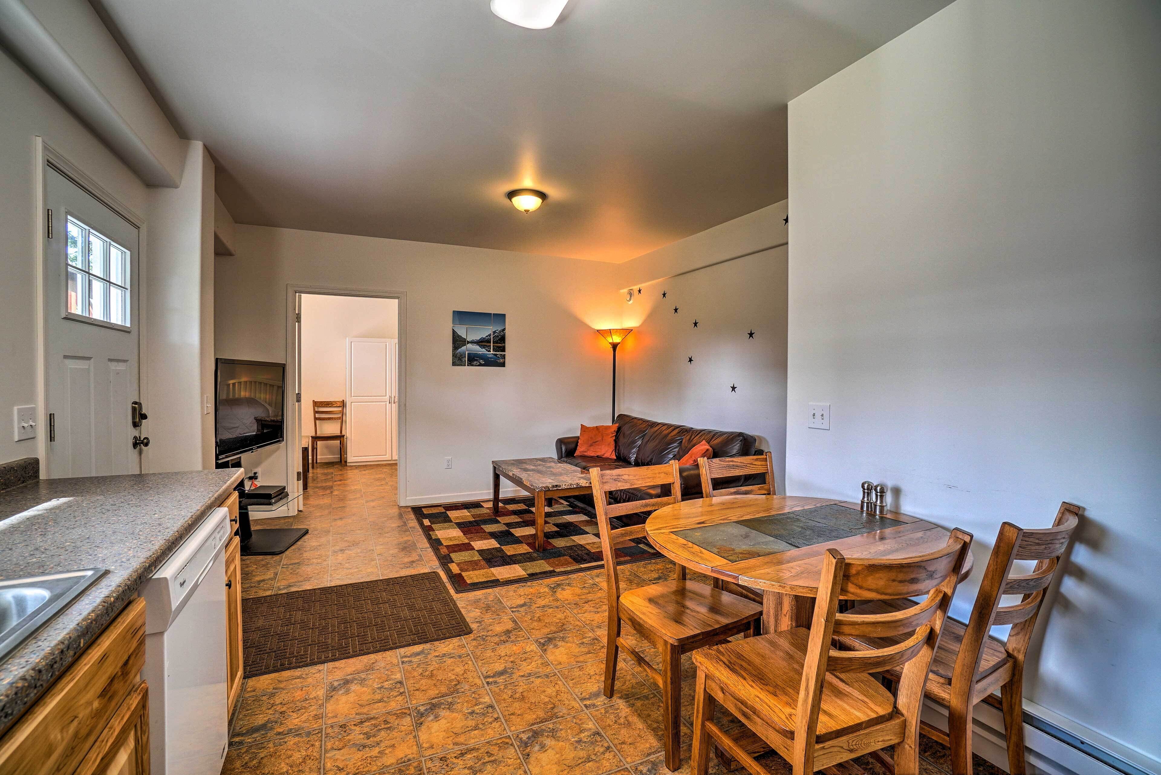 A compact kitchen with a dishwasher, a dining area with a wooden table and chairs, a sofa, and a checkered rug.