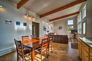 A spacious dining area with a wooden table and chairs, a kitchen with wooden cabinets, and a large window providing natural light. - Steps to Fritz Creek! 2-unit Family Retreat (Homer)
