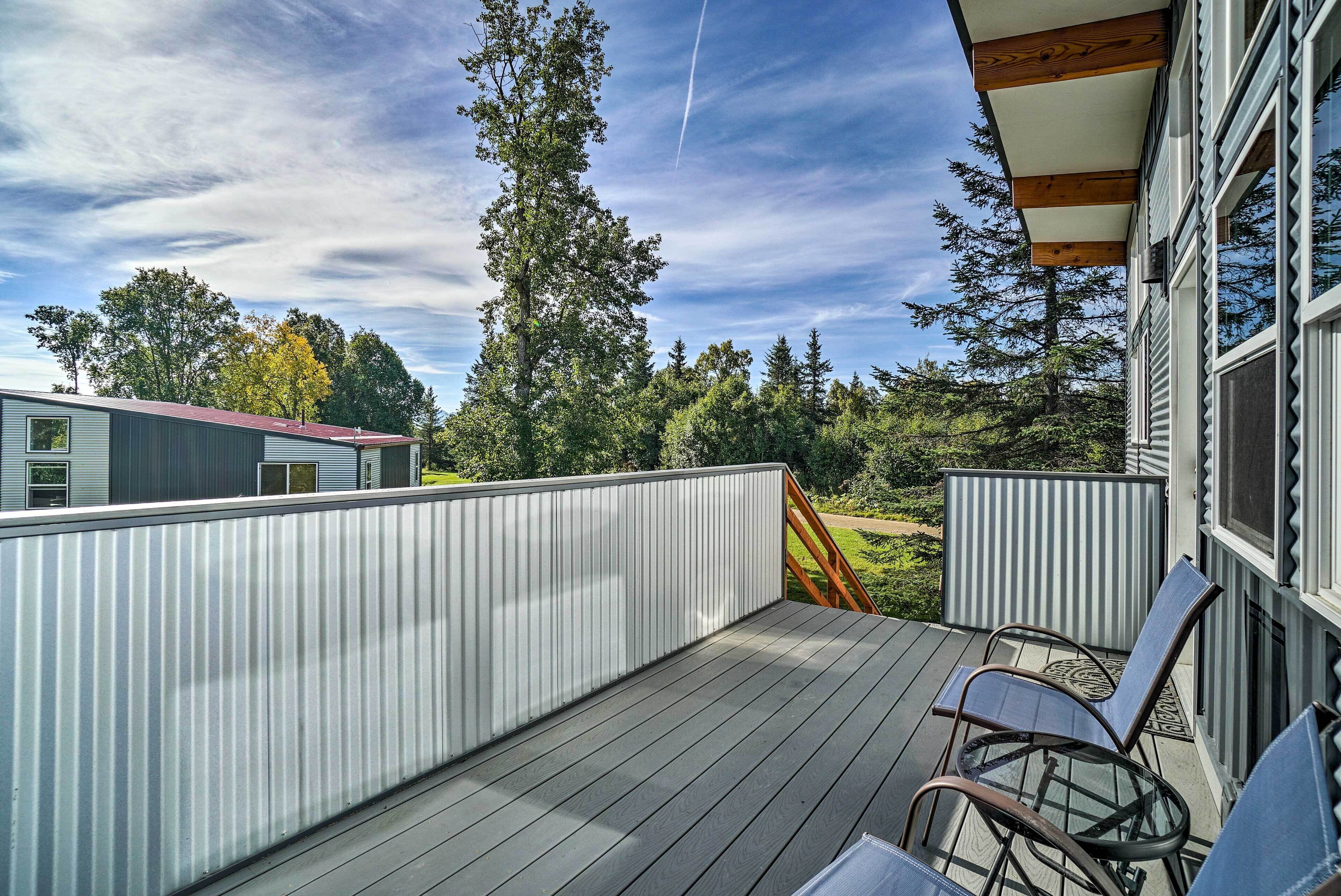 A wooden deck with metal railings, a glass door, and a view of trees and a building in the background.