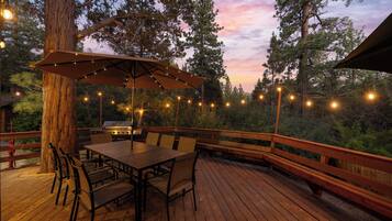 An outdoor deck with a wooden table and chairs, surrounded by trees and string lights.