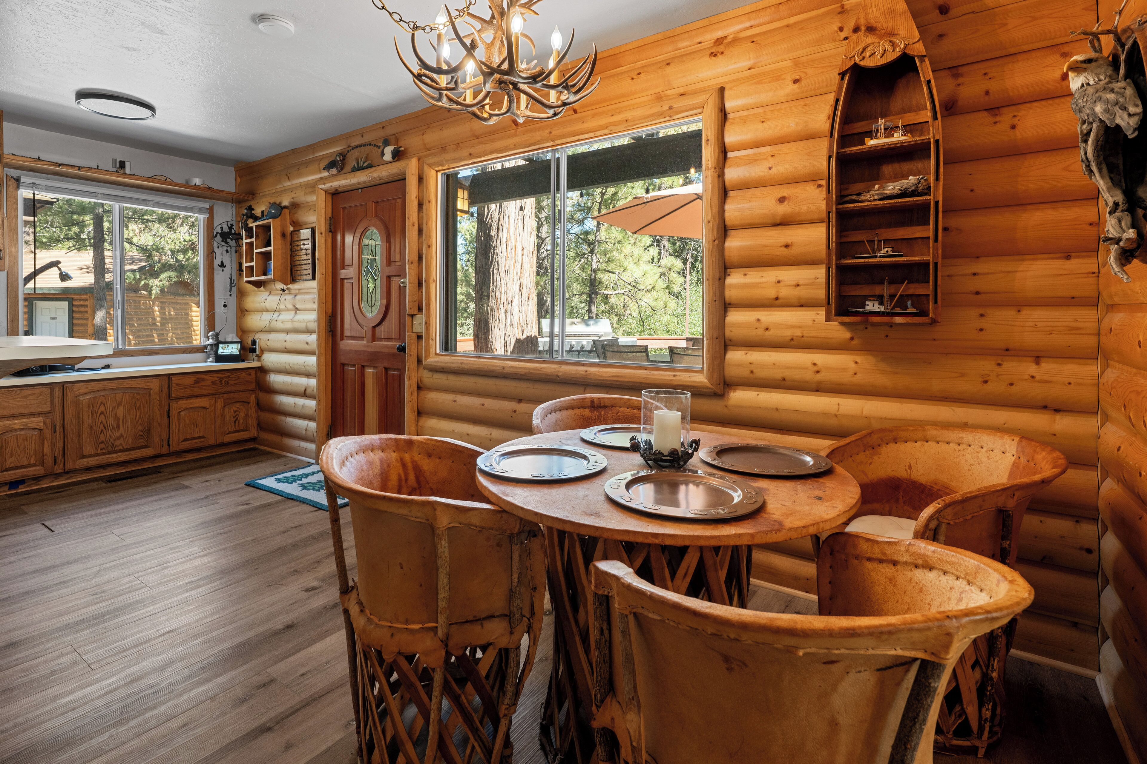 A wooden cabin interior with a dining table, chairs, a chandelier, and a window overlooking a forest.