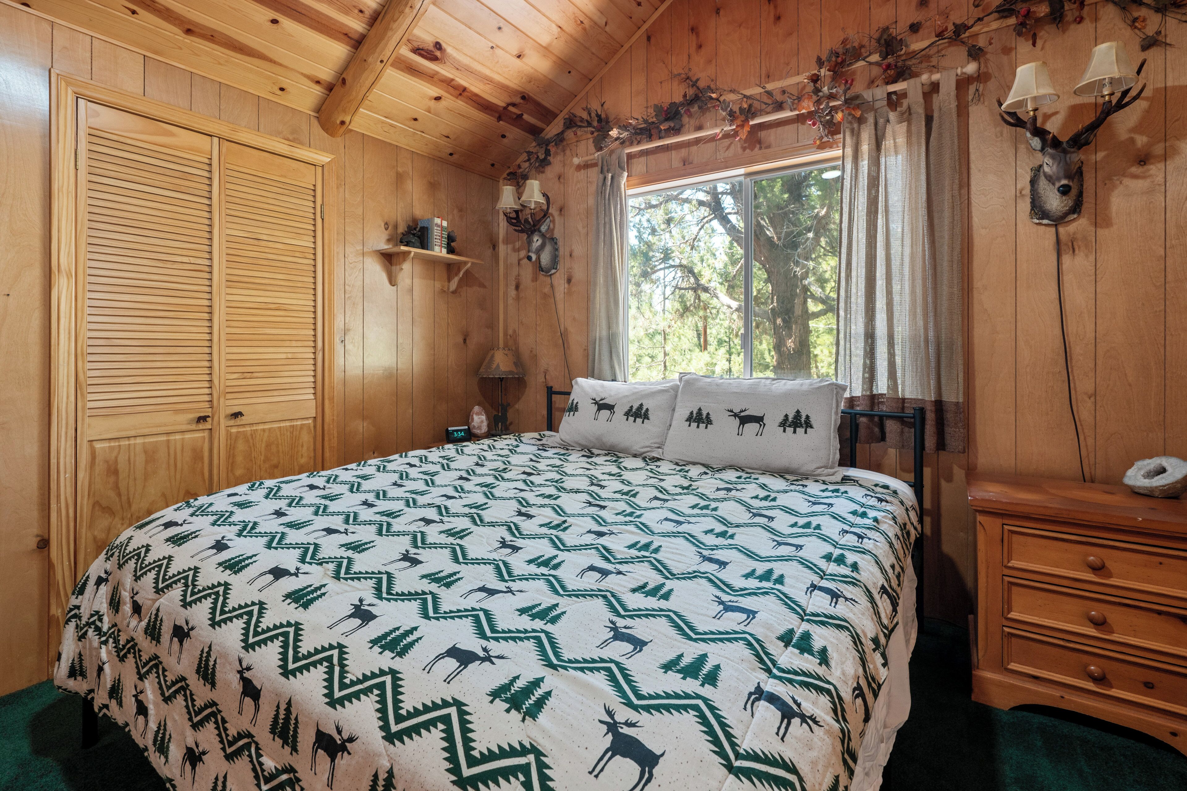 Un dormitorio de cabaña de madera con una cama, una mesita de noche, un aparador y una ventana con vistas a árboles.