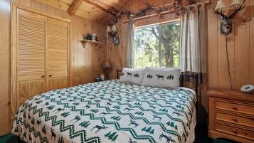Un dormitorio de cabaña de madera con una cama, una mesita de noche, un aparador y una ventana con vistas a árboles.