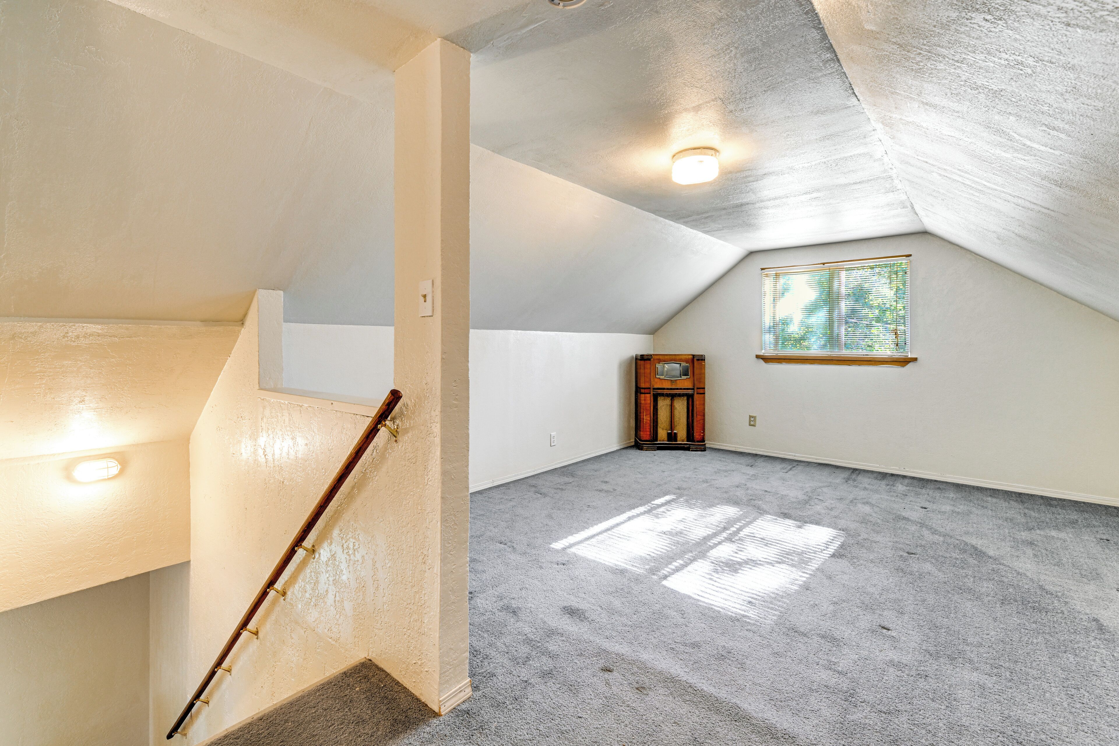 Amplia habitación en el ático con techo inclinado, una escalera de madera, un pequeño armario de madera y una ventana con persianas.