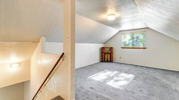 Amplia habitación en el ático con techo inclinado, una escalera de madera, un pequeño armario de madera y una ventana con persianas.