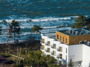 A modern building with a wooden facade and multiple balconies, located near a coastline with visible waves. - Navango - Beachfront, Sea View Pool & Sauna (Gąski)