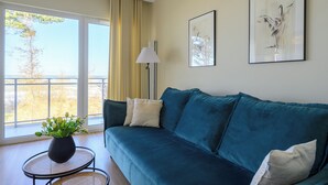 A living room with a teal sofa, a small round table with a plant, and a view through a large window. - Navango - Beachfront, Sea View Pool & Sauna (Gąski)