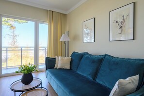 Uma sala de estar com um sofá verde-água, uma mesinha redonda com uma planta, e uma vista através de uma grande janela.