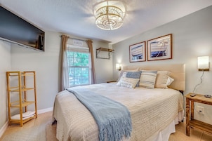 Quarto com cama, mesa de cabeceira, televisão fixada na parede, janela com cortinas e obra de arte na parede.